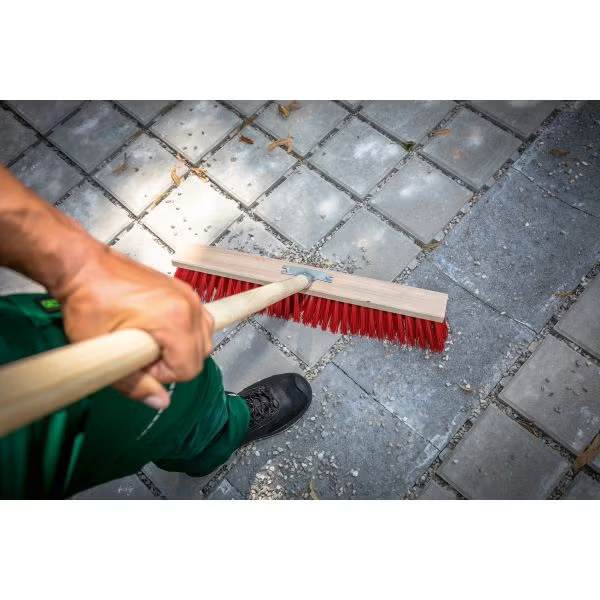 Eine Person kehrt mit einem Straßenbesen mit Holzstiel und Kunststoffborsten eine gepflasterte Fläche im Außenbereich.