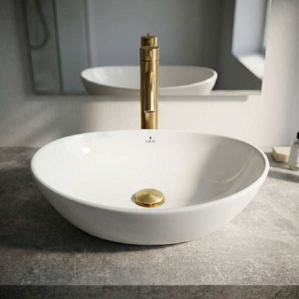 Ovales Aufsatzwaschbecken aus Keramik mit goldener Armatur und passendem Ablaufventil auf einer grauen Arbeitsplatte in Steinoptik.