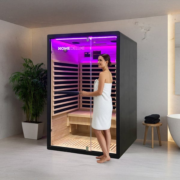Eine moderne Infrarotkabine von Home Deluxe mit Glasfront und Innenbeleuchtung in einem Badezimmer, neben einer Frau in einem Handtuch stehend.