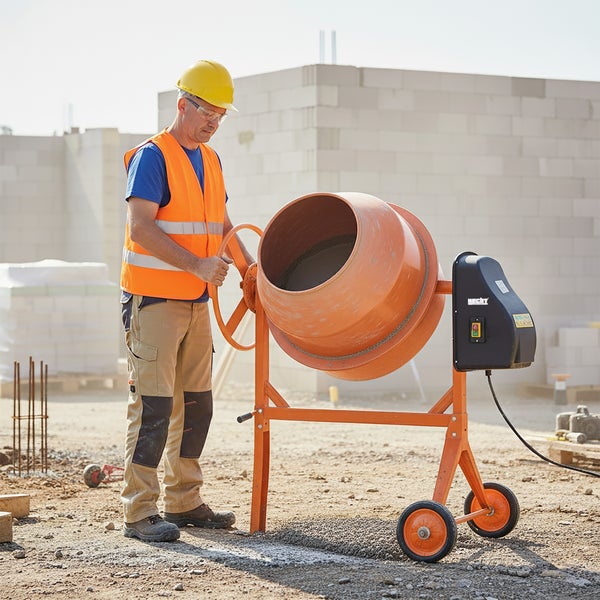 Bauarbeiter bedient einen orangefarbenen Hecht Betonmischer auf einer Baustelle. Er trägt Schutzkleidung wie Helm und Sicherheitsweste.