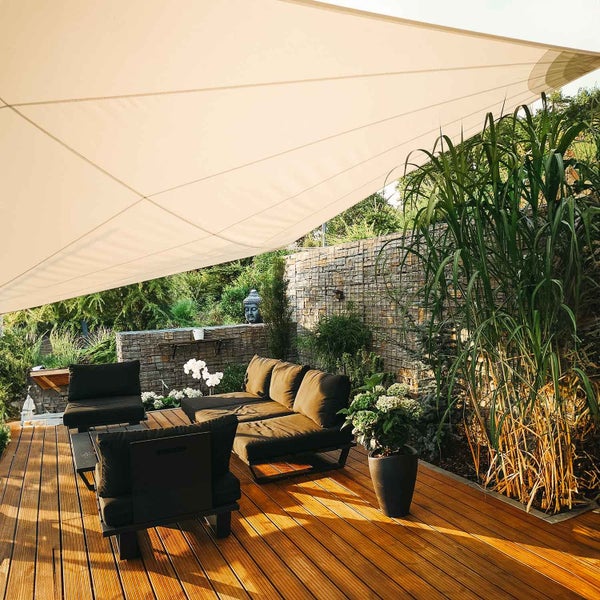 Großes helles Sonnensegel über einer modern eingerichteten Terrasse mit dunklen Loungemöbeln auf einem Holzdeck vor einer Steinwand.