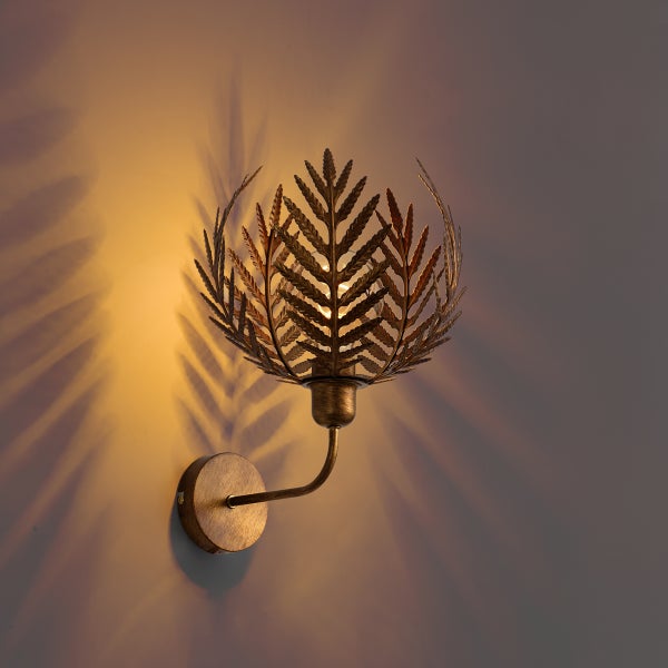 Wandleuchte aus Metall in Antik-Messing-Optik mit einem Schirm in Farnblatt-Design, der Schattenmuster an die Wand wirft.