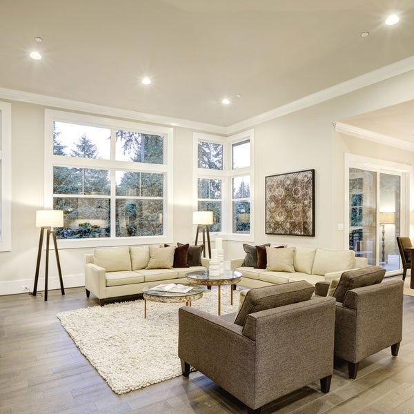 Helles Wohnzimmer mit zwei beigen Sofas, zwei grauen Sesseln, Glas-Couchtischen, Stativ-Stehlampen und großen Fenstern.