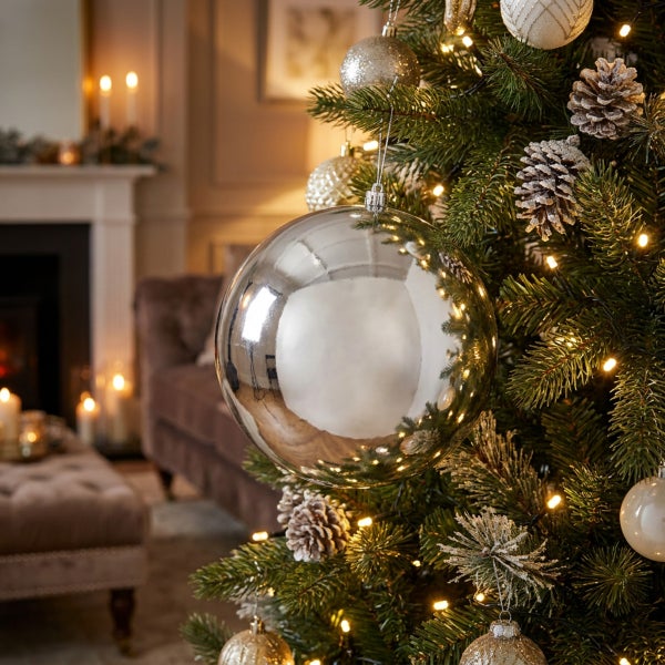 Eine glänzende, runde Christbaumkugel an einem geschmückten Weihnachtsbaum mit Tannenzapfen und Lichterkette in einem wohnlichen Raum.