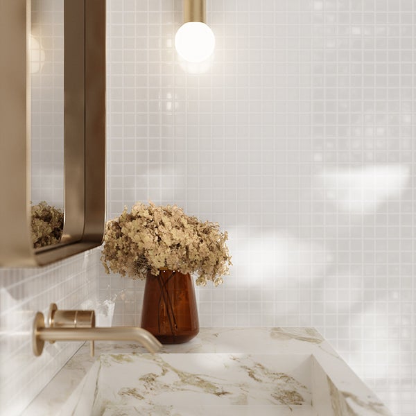 Modernes Badezimmer mit weißen Mosaikfliesen, Marmorwaschbecken, goldfarbener Armatur und Wandleuchte, wohnliche Atmosphäre mit Trockenblumen.
