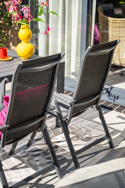 Zwei Gartenklappstühle mit dunklem Metallgestell und Netzbespannung an einem grauen Tisch auf einem gemusterten Außenteppich mit gelber Vase.