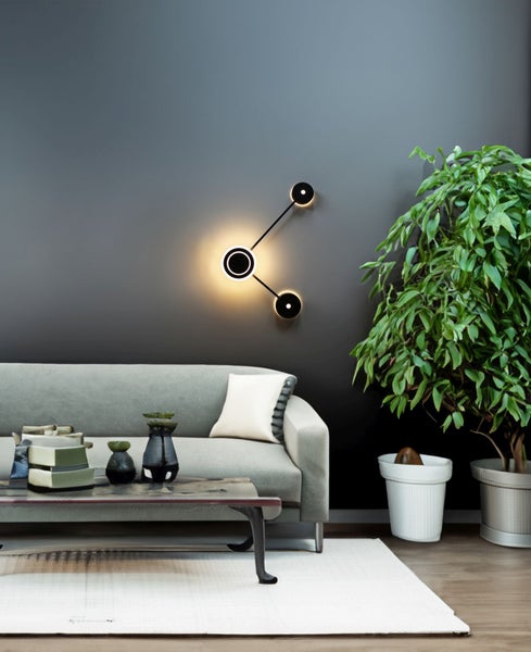 Moderne LED-Wandleuchte mit drei kreisförmigen Lichtelementen in einem Wohnzimmer mit Sofa und großer Zimmerpflanze an einer dunklen Wand.