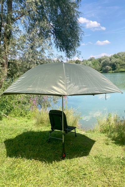 Großer grüner Anglerschirm mit schwarzem Angelstuhl am sonnigen Seeufer auf einer grünen Wiese unter Bäumen.