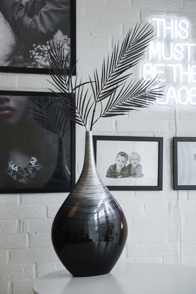 Bauchige Vase mit Silber-Schwarz-Verlauf und schwarzen künstlichen Palmwedeln vor einer weißen Ziegelwand mit Bilderrahmen.