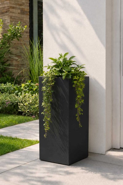 Moderner hoher rechteckiger Pflanzkübel mit hängenden Pflanzen auf einer Terrasse vor einer Hauswand mit Garten im Hintergrund