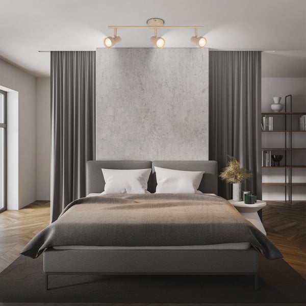 Modernes Schlafzimmer mit dreiflammigem Deckenstrahler über einem grauen Polsterbett, Betonwandpaneel und dunklen Vorhängen.