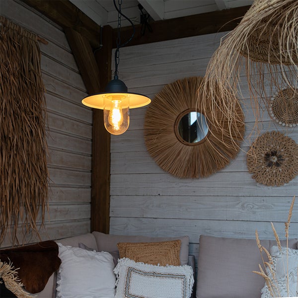Schwarze Pendelleuchte im Vintage-Stil mit Glaskolben in einer gemütlichen Sitzecke mit Holzterrasse und Boho-Wanddekoration aus Naturfasern.
