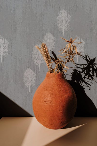 Eine bauchige Tonvase mit Trockenblumen vor einer Tapete mit baumartigem Muster.