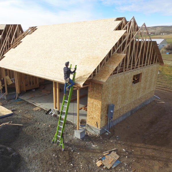 Person auf einer grünen Mehrzweckleiter arbeitet an einem Hausdach im Rohbau mit Holzkonstruktion und OSB-Platten.