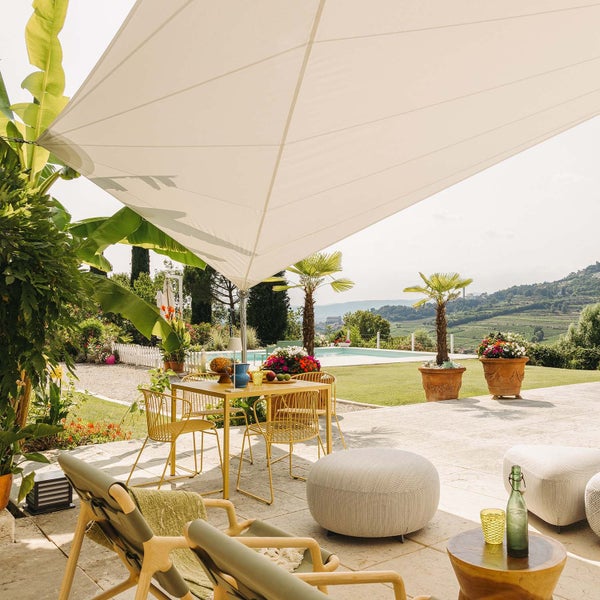 Sonnige Terrasse mit weißem Sonnensegel, gelber Essgruppe, gemütlichen Loungemöbeln und Blick auf einen Pool und Hügellandschaft.