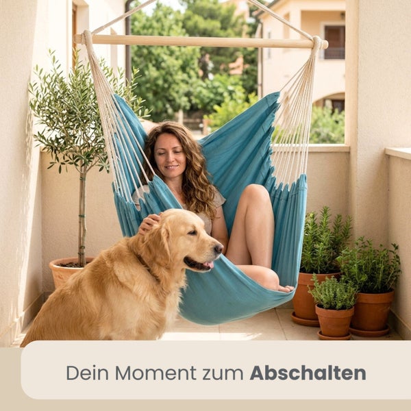 Frau sitzt in einem blauen Hängesessel aus Stoff auf einem Balkon und streichelt einen Hund. Topfpflanzen im Hintergrund.