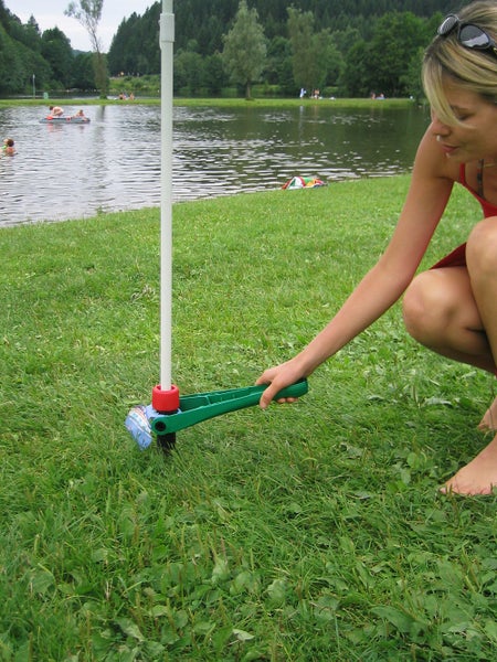 Eine Person schraubt einen grünen Bodenanker für Sonnenschirme mit einem Hebel in eine Rasenfläche an einem See.