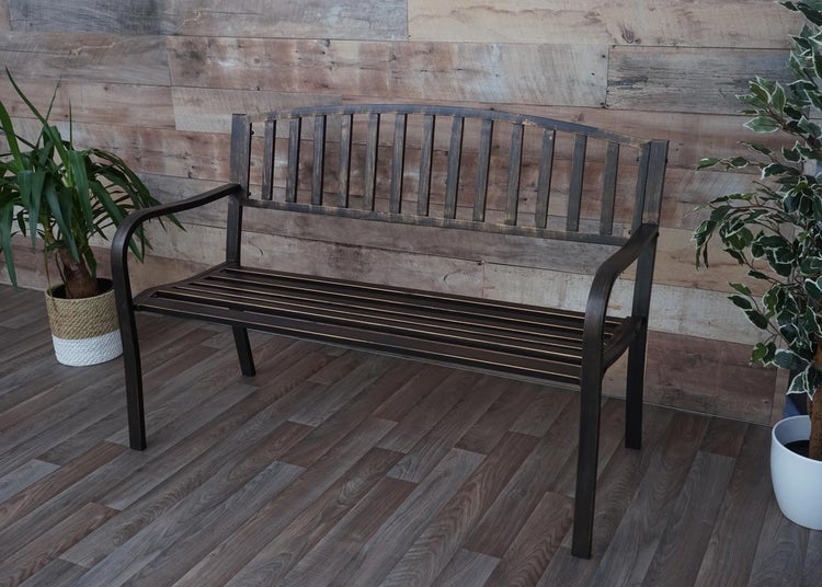 Metall-Gartenbank in Bronze-Optik mit vertikalen Rückenstreben und geschwungenen Armlehnen, zwischen zwei Zimmerpflanzen vor einer Holzwand.