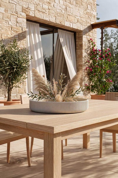 Holztisch für den Garten mit einer dekorativen Schale mit Pampasgras auf einer sonnigen Terrasse mit Steinwand und Olivenbaum.