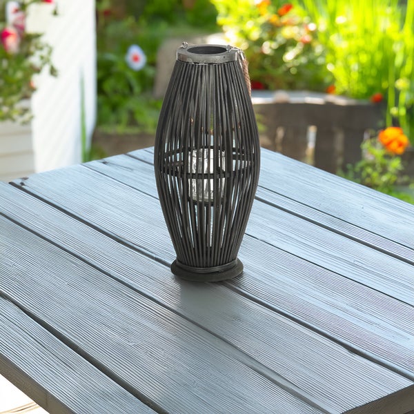 Ovale Windlicht-Laterne aus dunklem Bambusgeflecht mit Glaseinsatz auf einem grauen Gartentisch aus Holz.