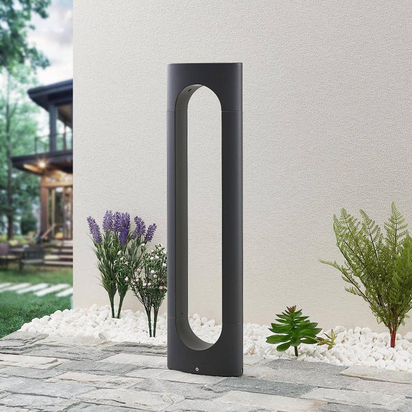 Moderne LED-Außenstandleuchte in Anthrazit mit ovaler Aussparung, platziert in einem Gartenbeet mit weißem Kies und Pflanzen.