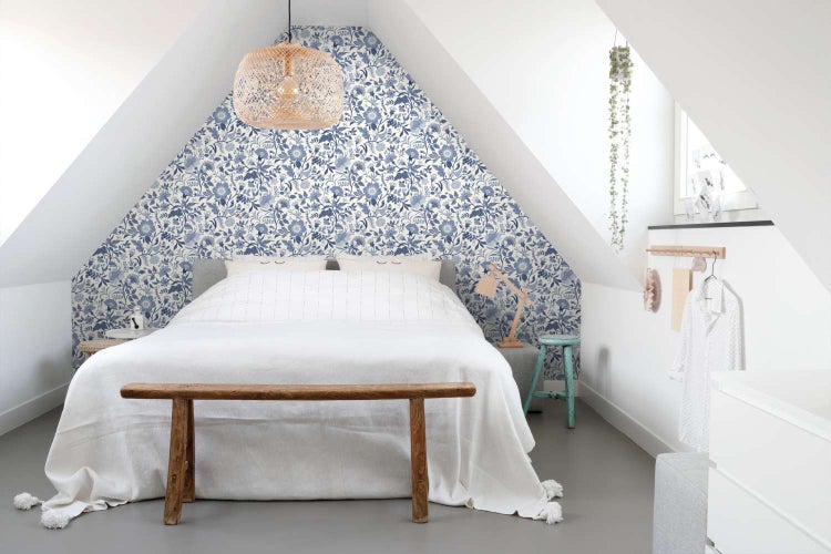 Helles Schlafzimmer unter dem Dach mit floraler Tapete hinter dem Bett und einer hölzernen Sitzbank am Fußende.