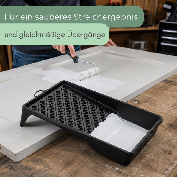 Eine Person trägt mit einer Farbrolle Farbe auf ein Werkstück auf, eine gefüllte Farbwanne steht auf dem Arbeitstisch bereit.