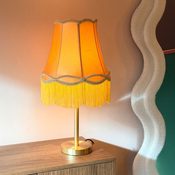 Tischleuchte mit Stoffschirm im Vintage Stil mit Fransen und metallischem Fuß auf einer Holzoberfläche.