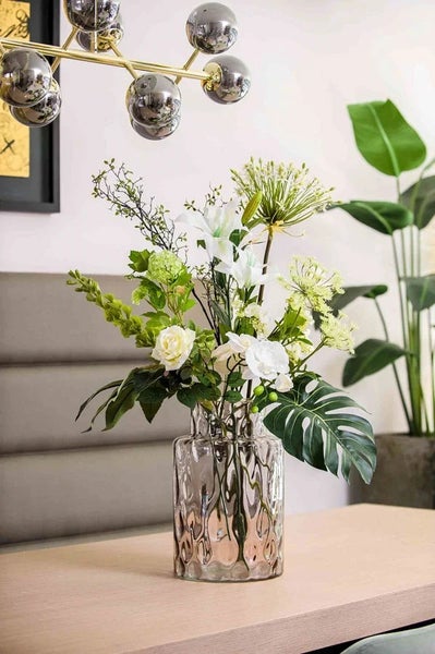 Künstlicher Blumenstrauß in strukturierter Glasvase auf Holztisch, moderne Raumdekoration mit weißen Lilien und Rosen.