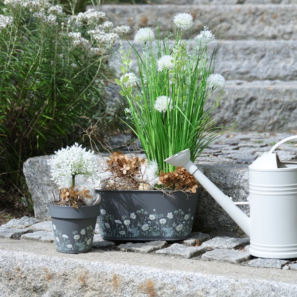 Graue Pflanztöpfe mit weißem Blumenmuster, Kunstpflanzen und eine weiße Gießkanne auf einer Steintreppe im Außenbereich.