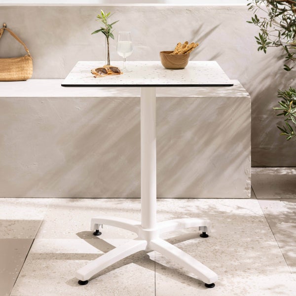Quadratischer weißer Bistrotisch mit Tischplatte in Terrazzo-Optik und zentralem Fußkreuz auf einer sonnigen Terrasse.