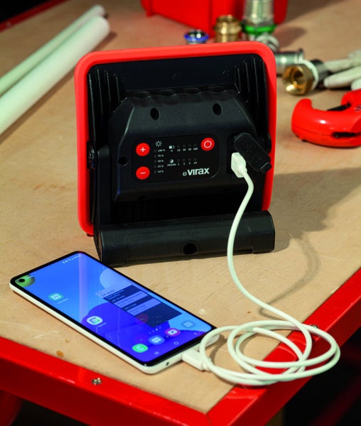 Virax Akku-Baustrahler mit Powerbank-Funktion lädt ein Smartphone über ein Ladekabel auf einer Werkbank mit Installationswerkzeugen.