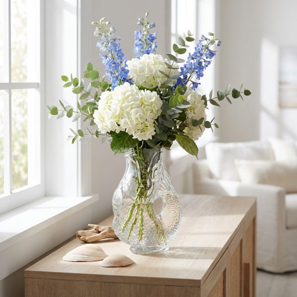 Blumenstrauß aus Hortensien und Rittersporn mit Eukalyptus in einer strukturierten Glasvase auf einem Holztisch vor einem Fenster