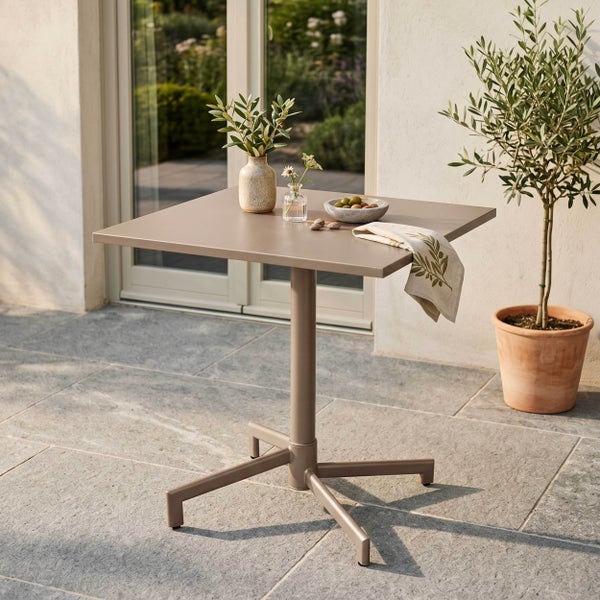 Quadratischer Gartentisch aus Metall auf einer Steinterrasse, dekoriert mit Vasen und Oliven, neben einem Olivenbaum in einem Tontopf.