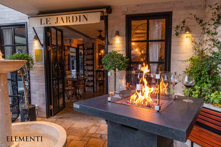 Elementi Gas-Feuerstelle als Tisch mit Glas-Windschutz und Flammen auf einer Terrasse mit Brunnen vor einem Restaurant. Elementi Logo.