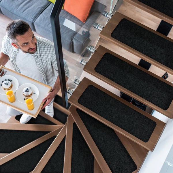 Mann trägt Frühstück auf einer Holztreppe mit textilen Stufenmatten und Glasgeländer in einem modernen Wohnraum.