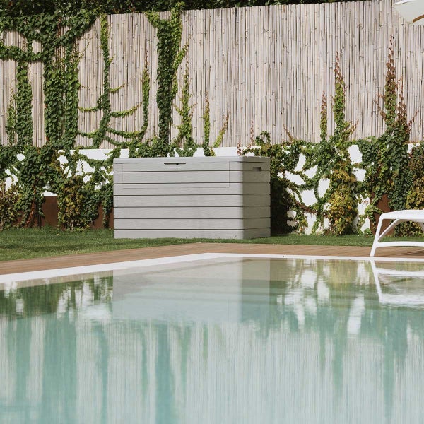 Gartenszene mit einer grauen Gerätebox aus Kunststoff, die neben einem Pool steht, vor einem mit Efeu bewachsenen Zaun