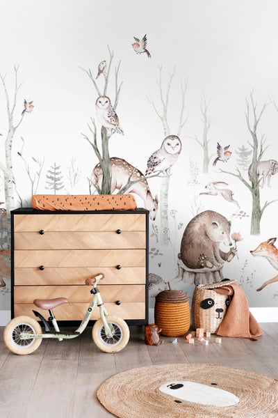 Kinderzimmer mit Kommode, Laufrad, Körben und einem Teppich mit Tiermotiv vor einer Waldtapete