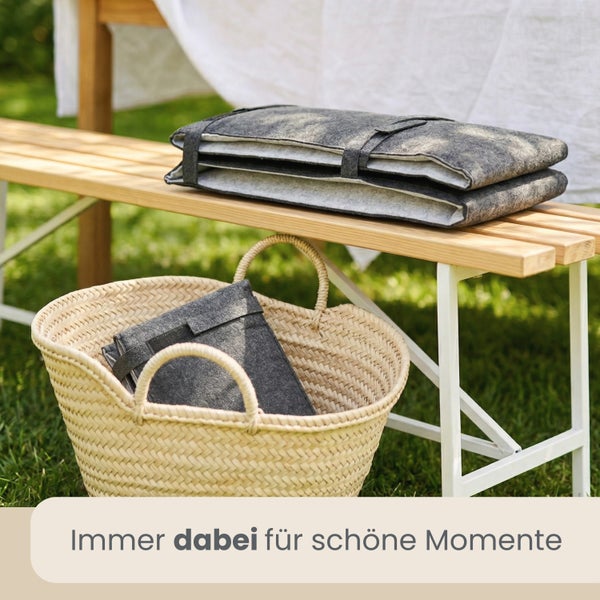Zusammengeklappte Sitzkissen aus Filz auf einer Holzbank mit einem geflochtenen Korb im Garten.