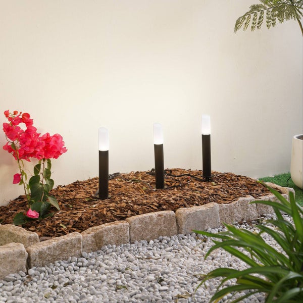 Drei schwarze zylindrische LED-Außenleuchten mit weißem Licht in einem Gartenbeet mit Rindenmulch und Steineinfassung.