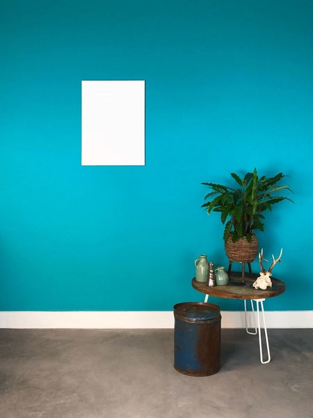 Modern eingerichteter Raum mit türkisfarbener Wand, weißem Bilderrahmen, rundem Holztisch mit Metallbeinen, Zimmerpflanze und Betonboden.