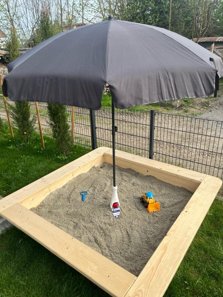 Quadratischer Sandkasten aus Holz mit Sand, einem grauen Sonnenschirm in der Mitte und Spielzeugbagger in einer Gartenanlage.