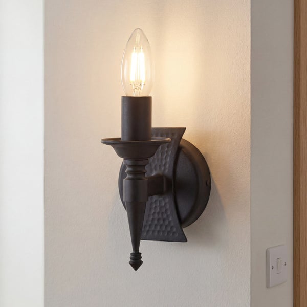 Rustikale Wandleuchte aus dunklem Metall mit gehämmerter Rückplatte und einer leuchtenden LED-Kerzenlampe.