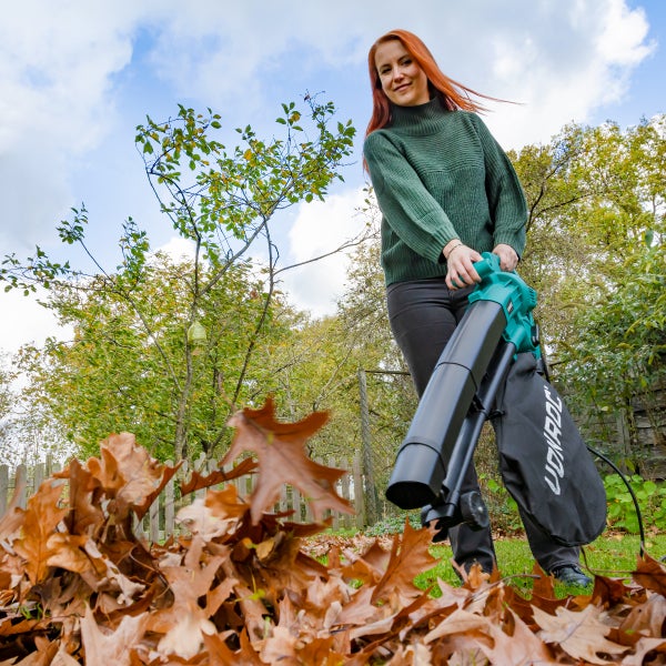 Eine Frau nutzt einen elektrischen Laubbläser mit Fangsack im Garten, um herbstliches Laub zusammenzutragen.