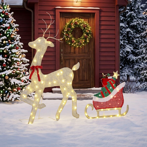 Beleuchtete Weihnachtsdekoration bestehend aus einem Rentier und einem Schlitten vor einem Haus mit Weihnachtsbaum und Adventskranz.