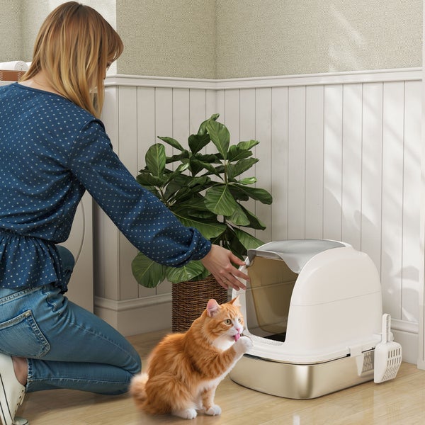 Eine Frau kniet neben einer modernen Haubentoilette für Katzen in einem hellen Raum, eine orangefarbene Katze sitzt davor.