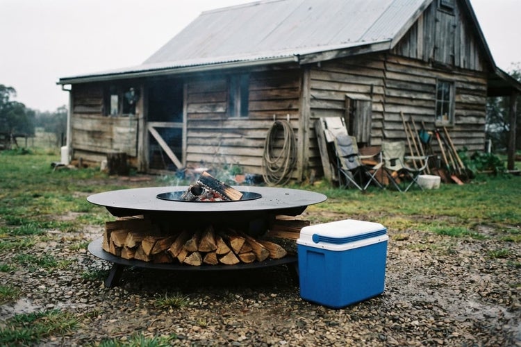 Feuerschale mit Grillplatte auf einem ländlichen Anwesen vor einer Holzhütte mit gestapeltem Brennholz und einer Kühlbox im Garten.