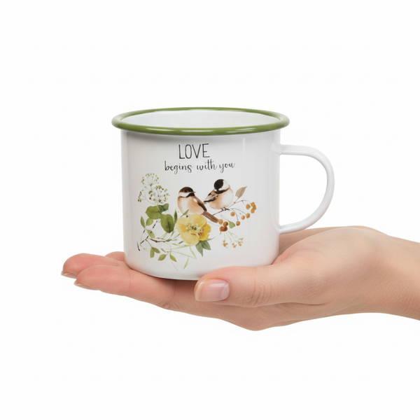 Weißer Becher im Emaille-Stil mit grünem Rand, Vogel- und Blumenmotiv sowie Text LOVE begins with you, in einer Hand gehalten.