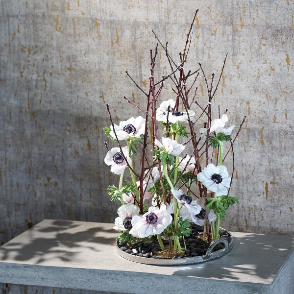 Modernes Gesteck mit weißen Anemonen, braunen Zweigen und schwarzen Kieselsteinen in einer silbernen Schale auf einer Steinfläche.