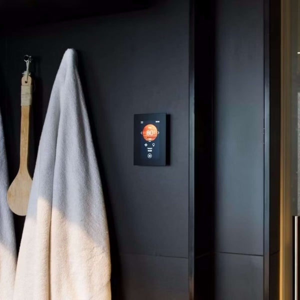 Digitale Saunasteuerung mit Touchdisplay montiert an einer Wand neben einem Handtuch und einer Saunakelle.